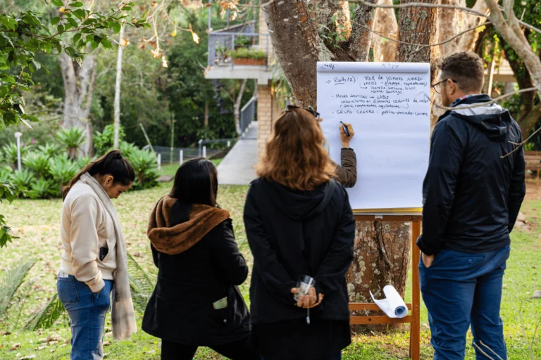 Four people stand outdoors by a whiteboard attached to a tree, with one writing while the others watch.